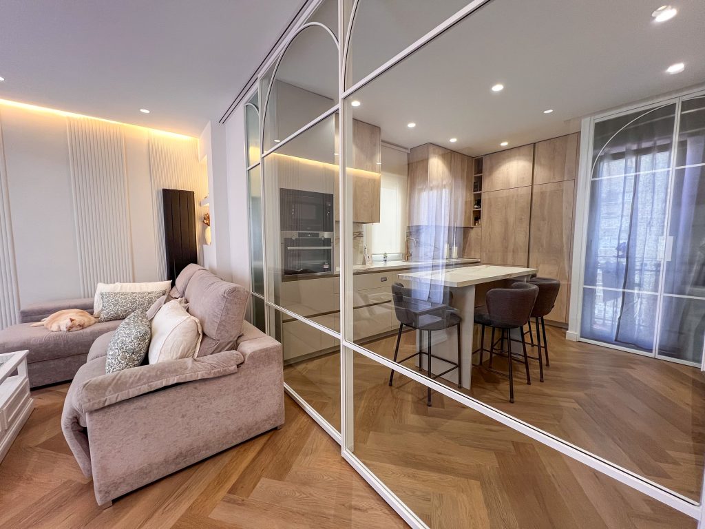 Cocina moderna de madera con isla central, separada del salón mediante cerramiento acristalado con perfiles blancos, en vivienda de diseño contemporáneo.